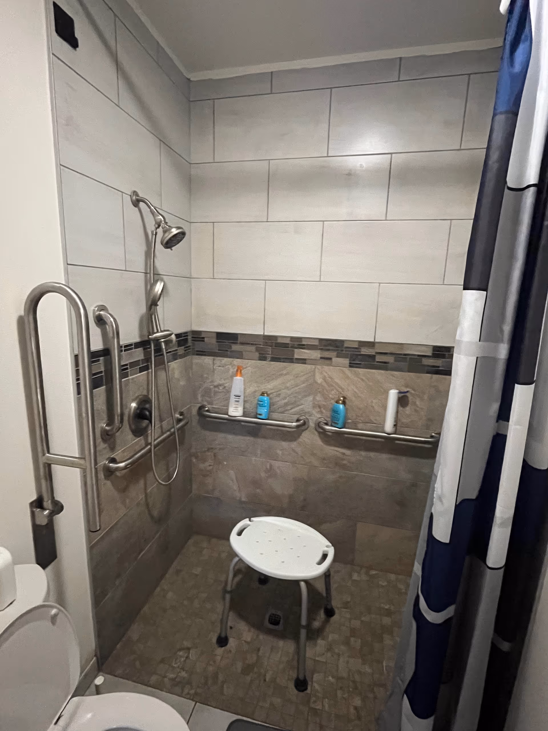 Accessible bathroom with shower, grab bars, a shower chair, and toiletries on a tiled wall.
