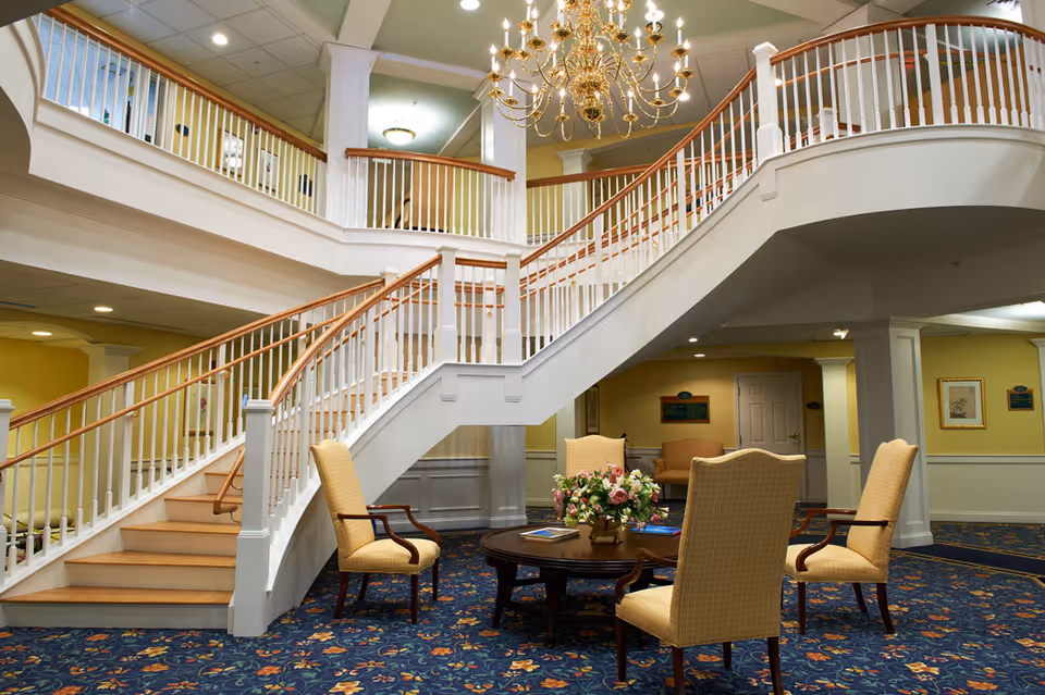 Interior view of a senior living facility lobby area featuring a grand staircase with white railings and wooden handrails. Below the staircase is a seating area with four upholstered chairs around a round wooden table decorated with a floral arrangement. The carpet has a floral pattern, and a large chandelier hangs from the ceiling. The walls are painted yellow with white trim.