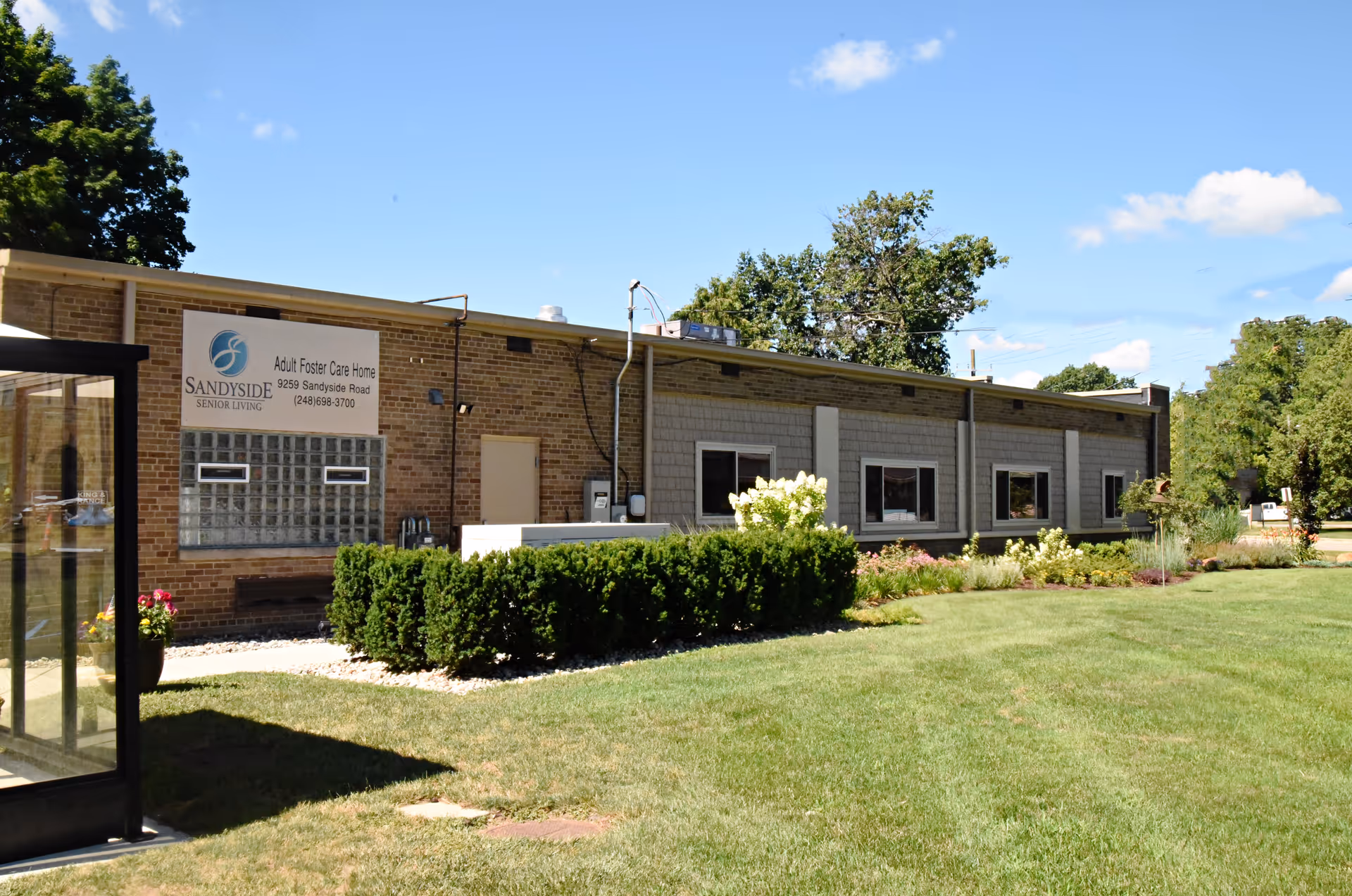 Exterior view of Sandyside Senior Living, a single-story brick building with several windows, surrounded by green grass, bushes, and trees under a clear blue sky.