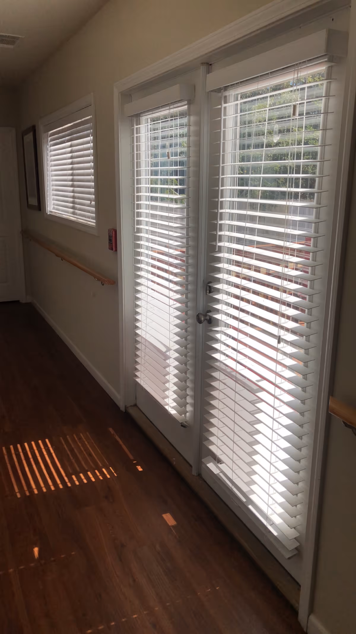 A hallway with wooden flooring and white walls featuring a handrail on the left side. There are two windows with white blinds on the left wall and a set of double glass doors with white blinds on the right side, allowing sunlight to cast shadows on the floor.