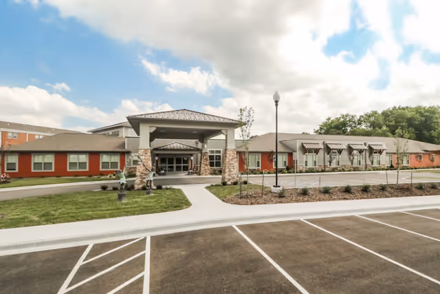 Exterior view of Carnegie Village Senior Living Community showing a single-story building with a covered entrance supported by stone pillars, a parking lot in the foreground, and a partly cloudy sky above.