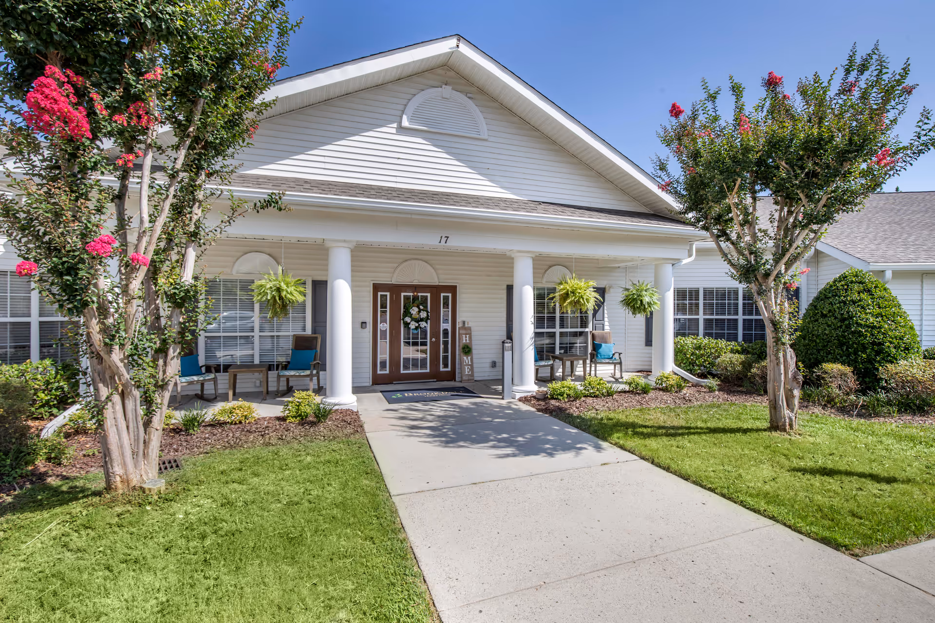 Front entrance of a senior living facility with white siding, a covered porch supported by white columns, two hanging plants, and outdoor seating with chairs and small tables. There are flowering trees and well-maintained landscaping on either side of the concrete walkway leading to the double glass doors.