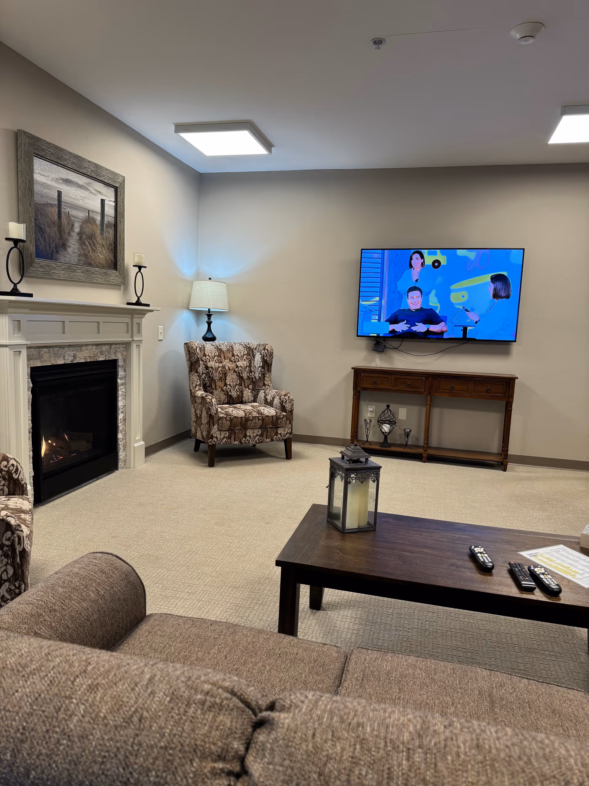 Living room in a senior facility featuring a fireplace, patterned chairs, sofa, coffee table, and TV.