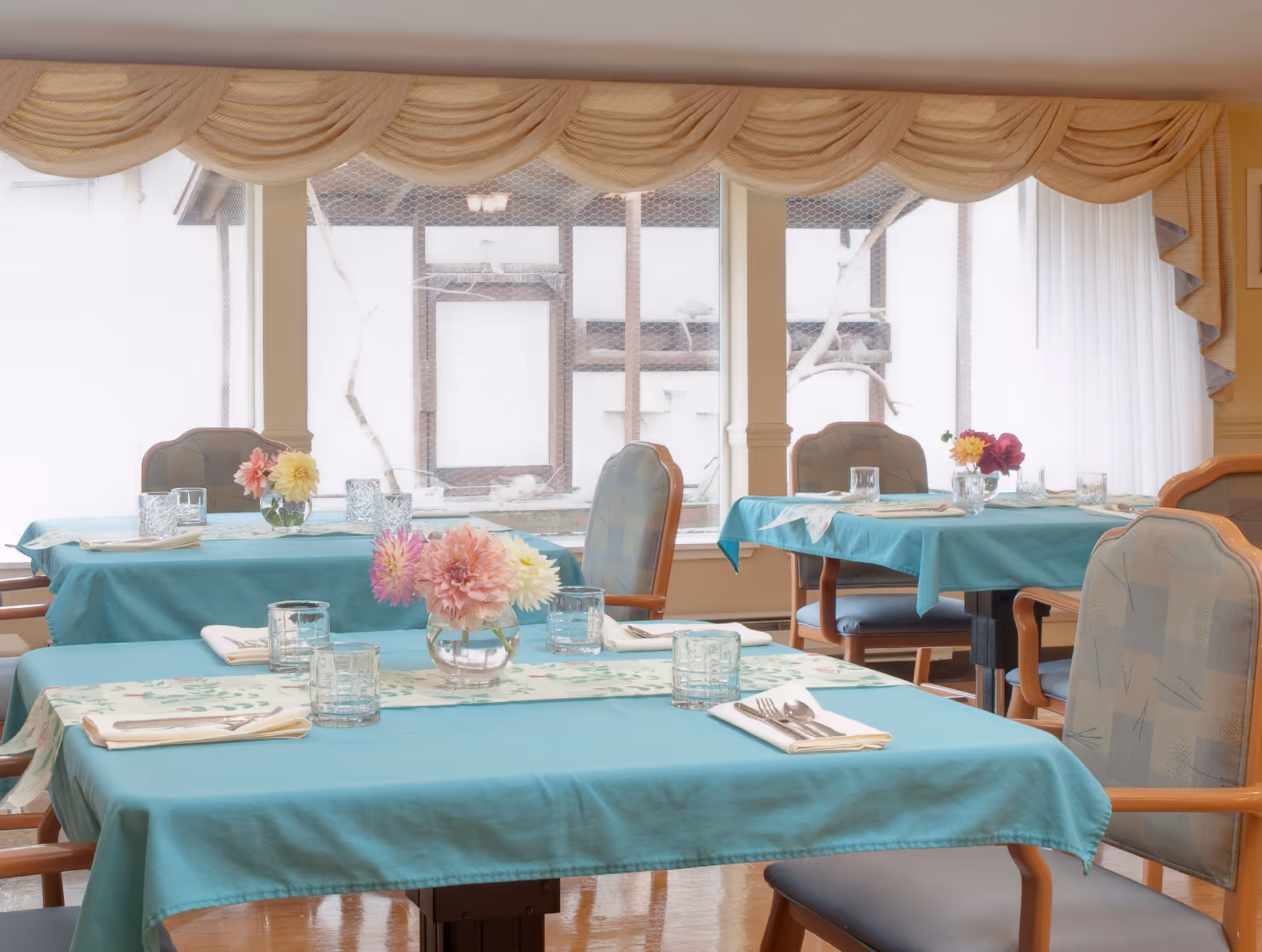 Dining room with tables covered in teal tablecloths, each set with glassware, napkins, silverware, and small vases of colorful flowers. Large windows with beige valances and white curtains allow natural light to fill the room.