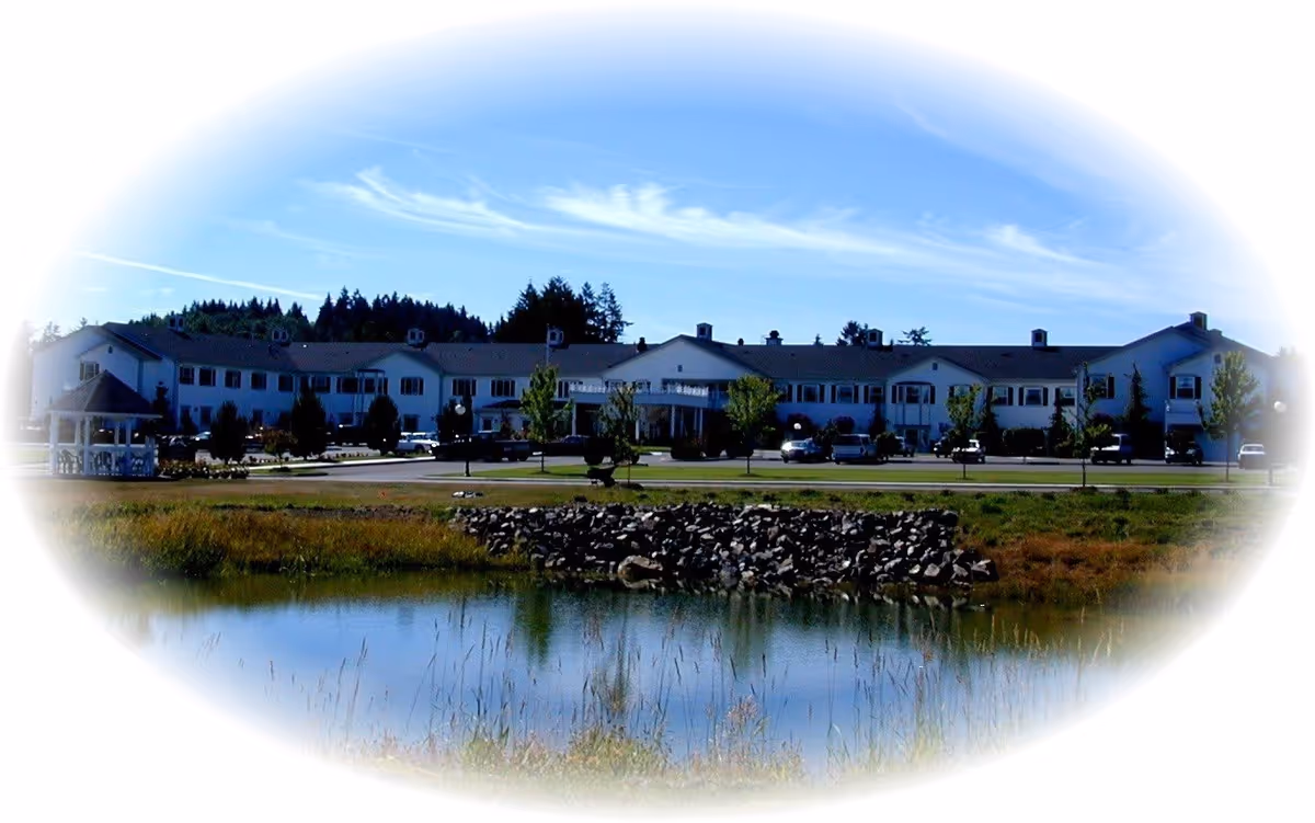 Exterior view of a large two-story residential building with multiple windows and a central entrance, surrounded by a parking area with cars, trees, and a small pond with rocks in the foreground under a blue sky with wispy clouds.