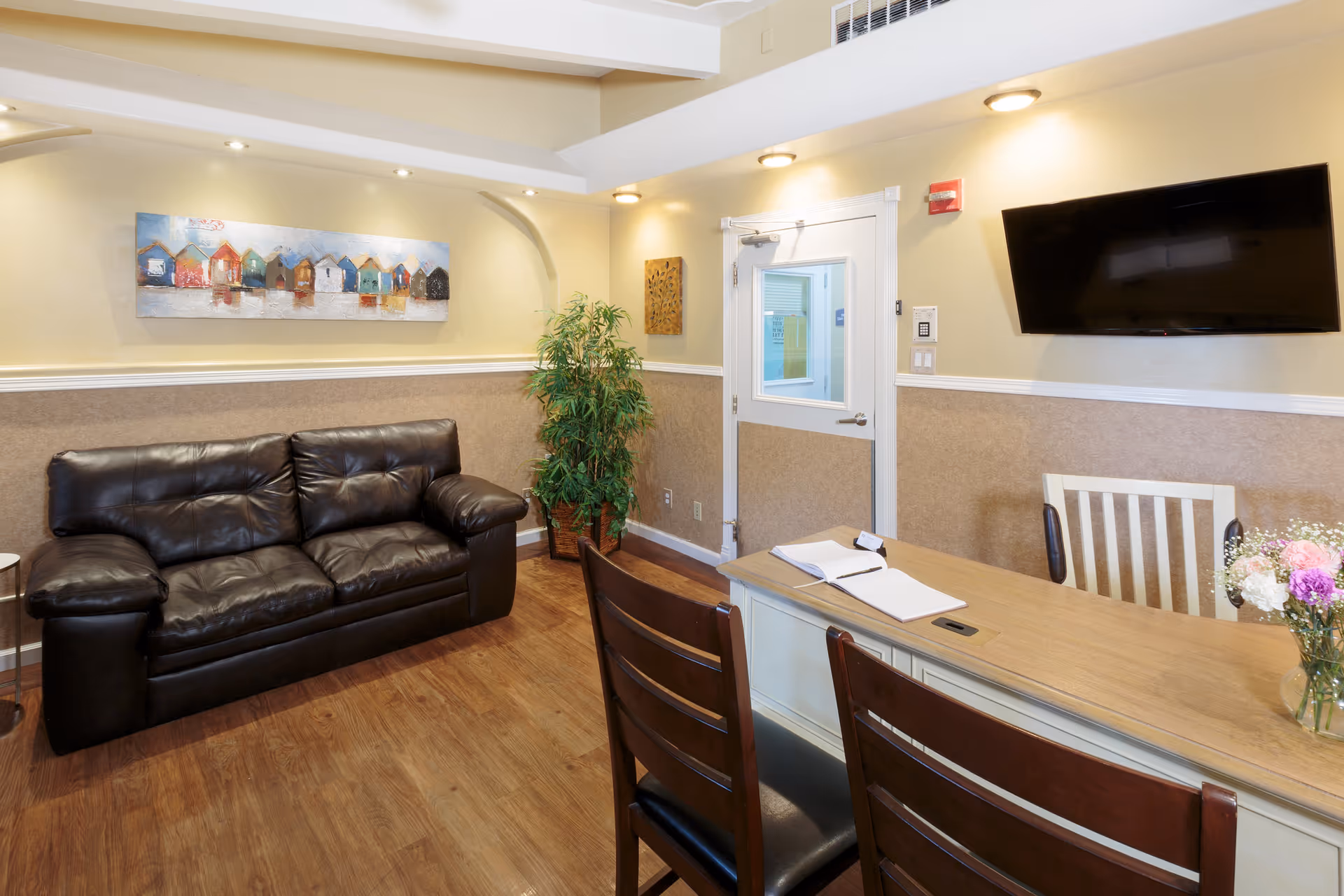 Cozy common room with a leather sofa, table and chairs, wall-mounted TV, artwork, and a potted plant.
