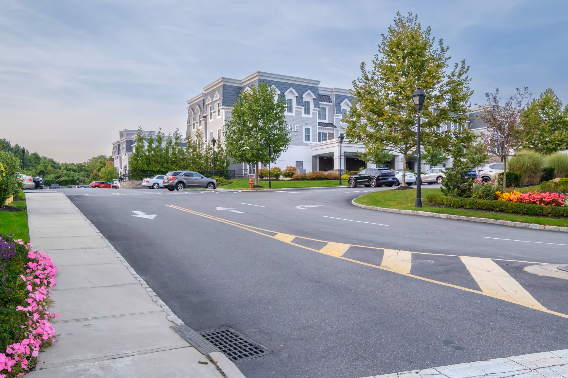 Exterior view of Encore Luxury Living facility showing a paved driveway with directional arrows, parked cars, landscaped flower beds, trees, and a multi-story building with gray siding and white trim under a partly cloudy sky.