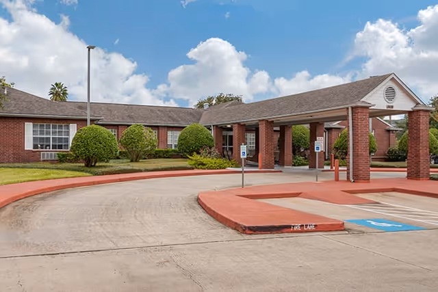 Exterior view of a single-story brick building with a covered entrance supported by brick columns. The driveway curves around the entrance with marked parking spaces, including handicapped spots. Well-maintained bushes and greenery surround the building under a partly cloudy sky.