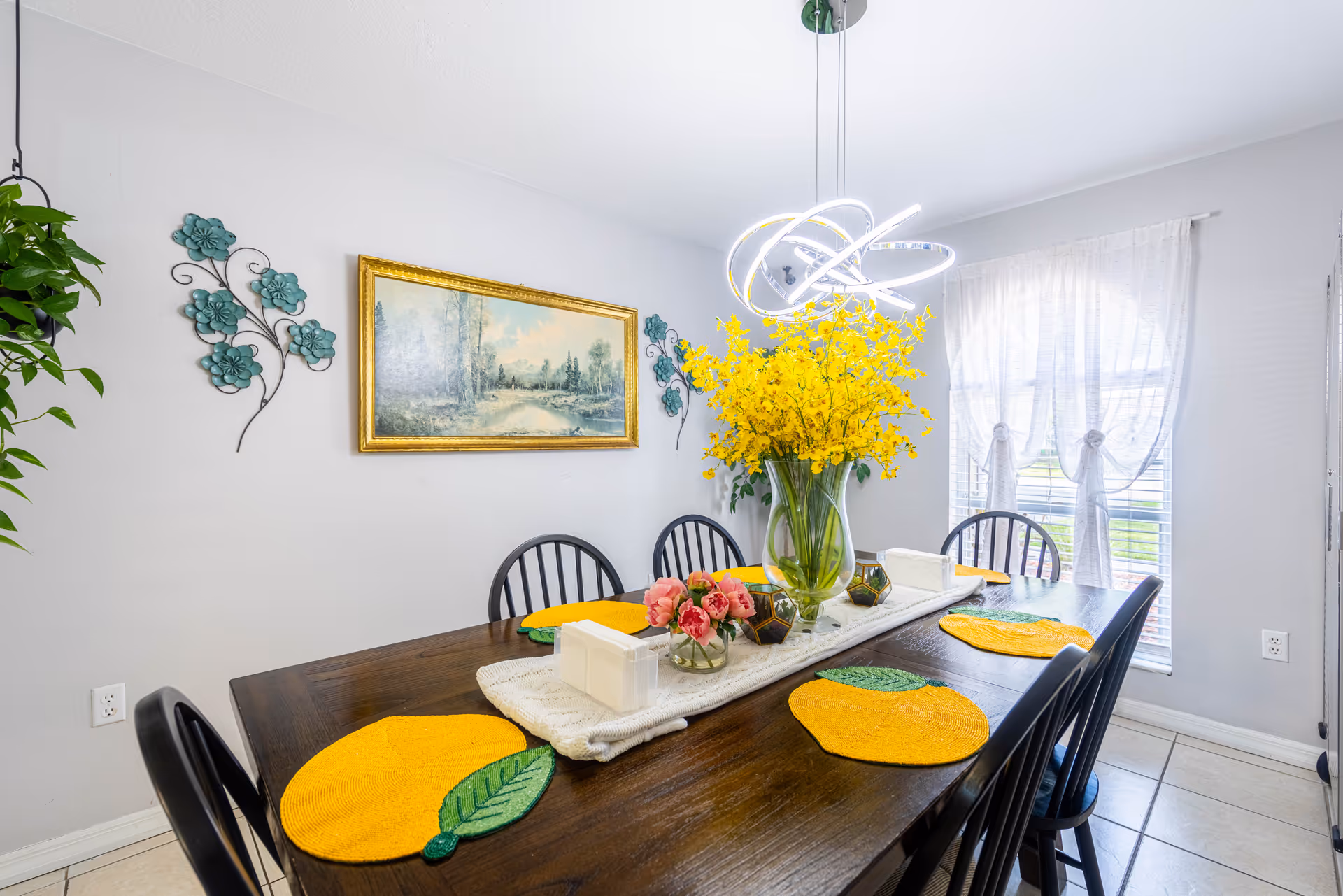 Bright dining room with a long wooden table, yellow flower centerpiece, and sheer-curtained window.