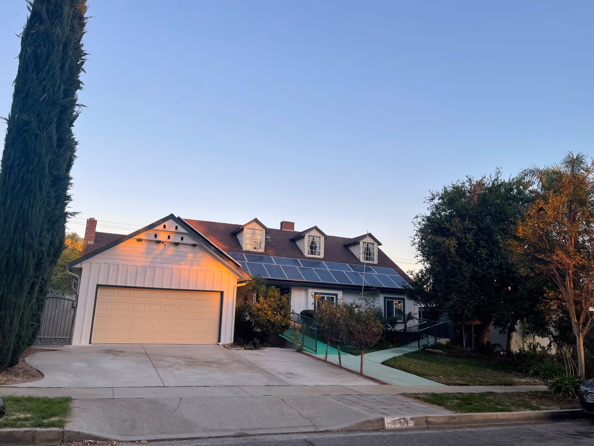 Residential house with a two-car garage, solar panels, trees, and a sidewalk.