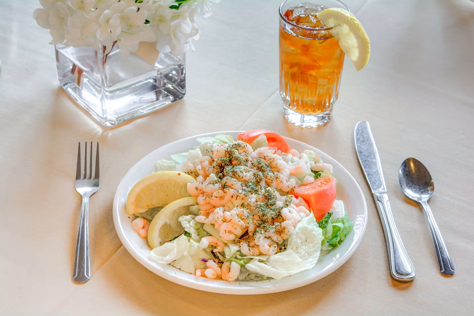 A plate of shrimp salad with lettuce, tomato slices, lemon wedges, and herbs on a white tablecloth. Next to the plate are a fork, knife, and spoon, and a glass of iced tea with a lemon wedge. A clear vase with white flowers is also on the table.