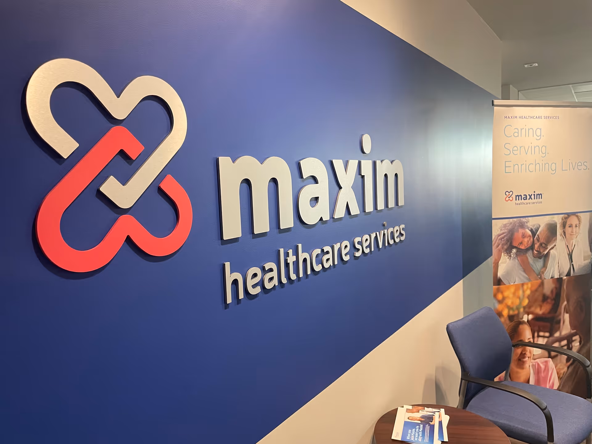 Blue wall with Maxim Healthcare Services logo, banner, and seating in an interior lobby.