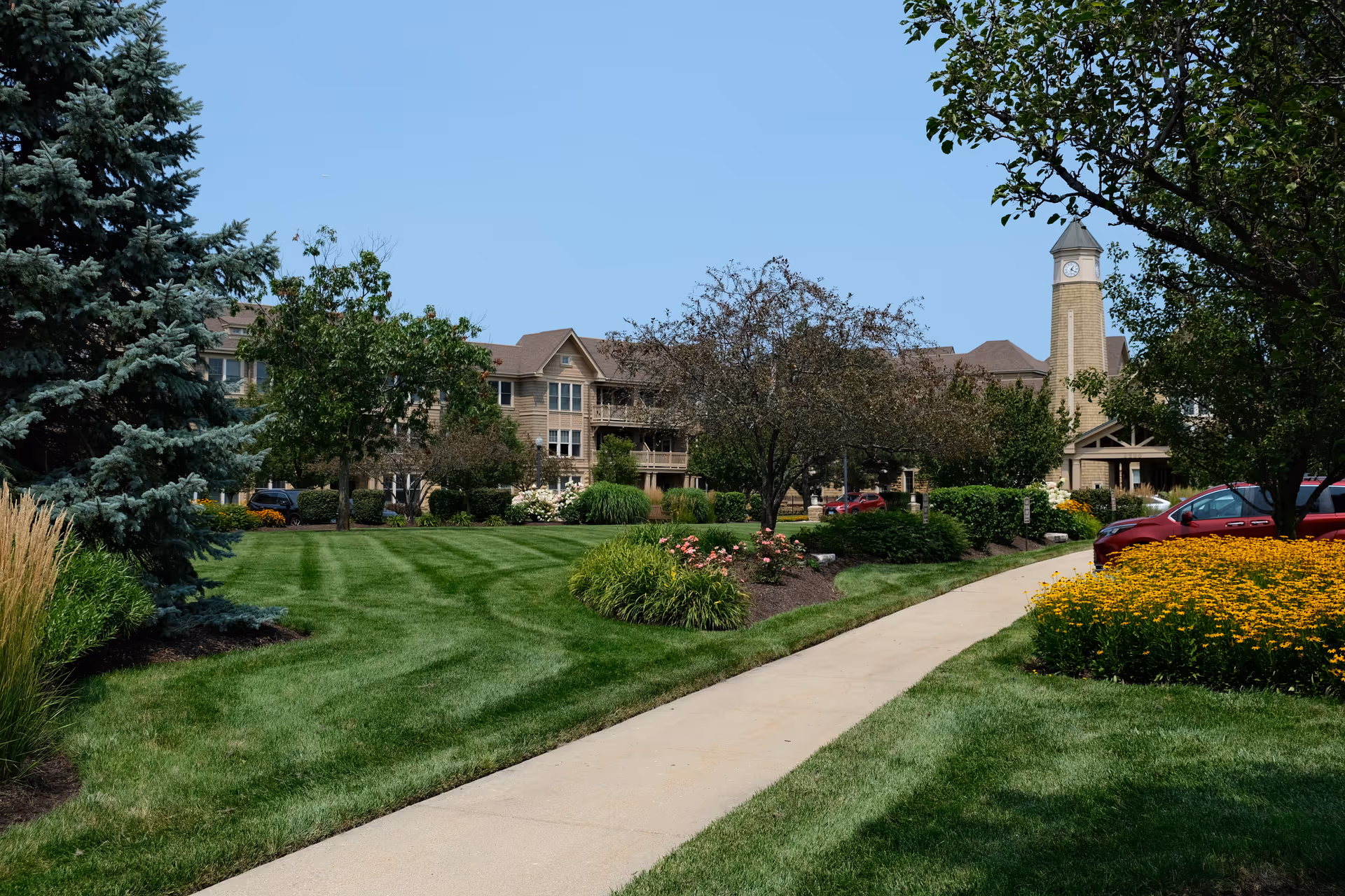 A landscaped sidewalk leads through manicured lawns and flower beds to a multi-story senior living building with a clock tower.