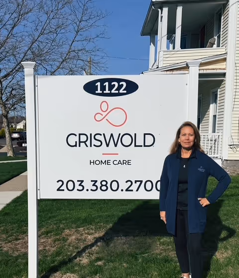 A Griswold Home Care sign on a lawn with a woman standing beside it outside.
