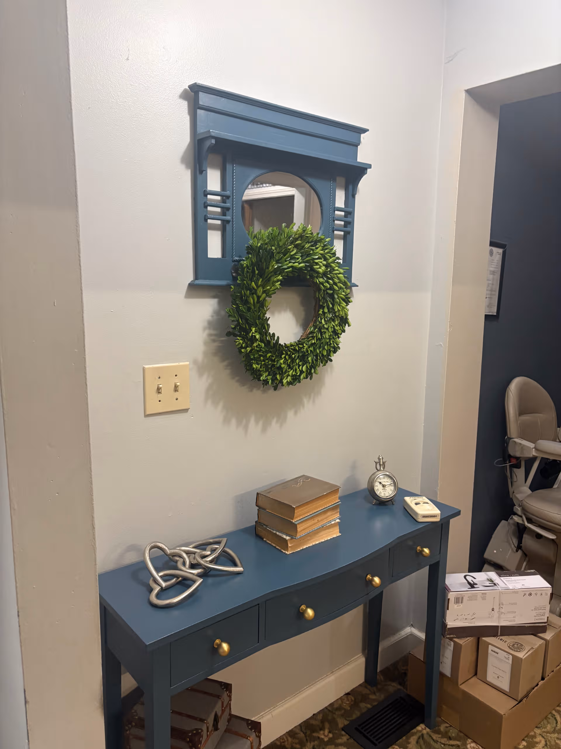 Blue console table against a light wall with a wreath, mirror, clock, books, and boxes.