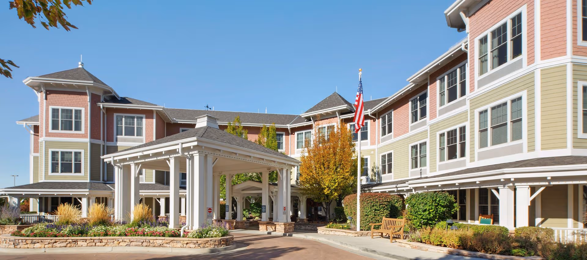 Exterior view of Sunrise at Holladay senior living facility showing a multi-story building with a covered entrance, landscaped garden beds, an American flag on a flagpole, and benches along the walkway under a clear blue sky.