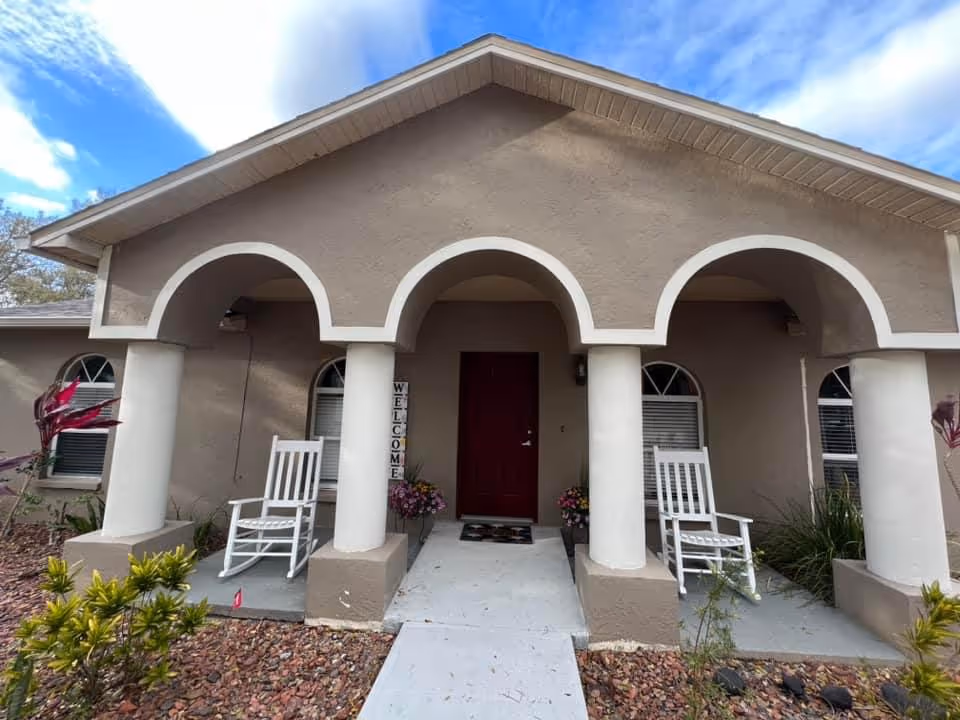 Tan building front with arched porch, two white rocking chairs, and a red front door.