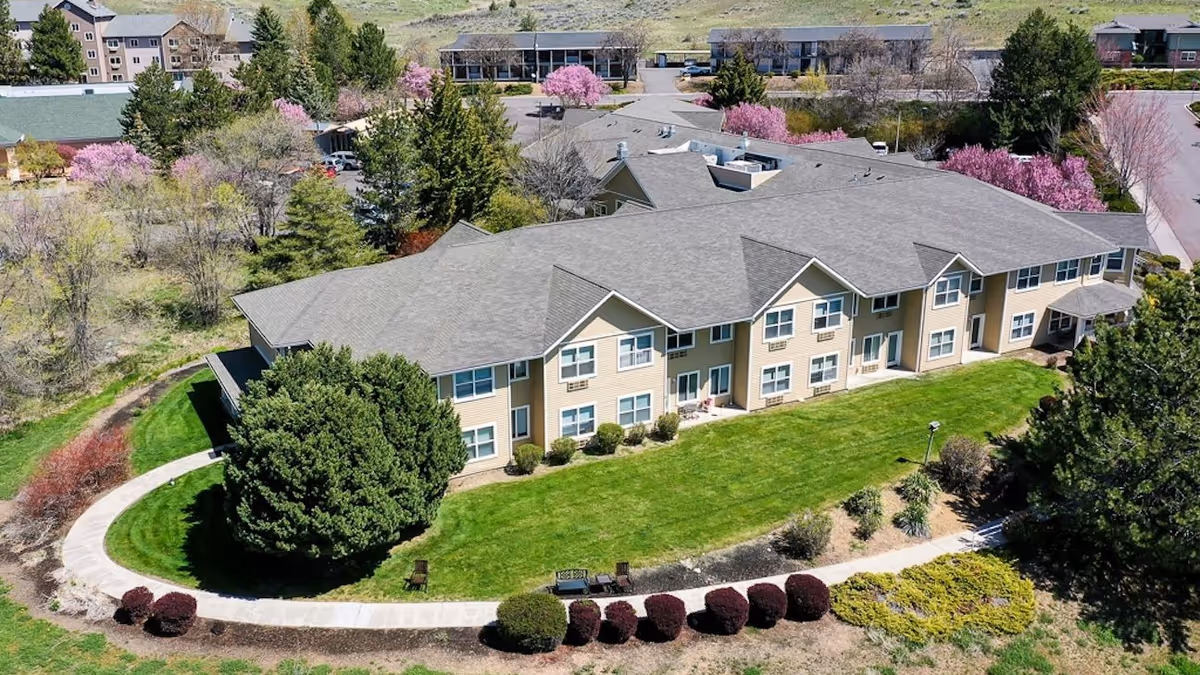 A beige senior living facility with curved walkway, manicured lawn, and surrounding trees.