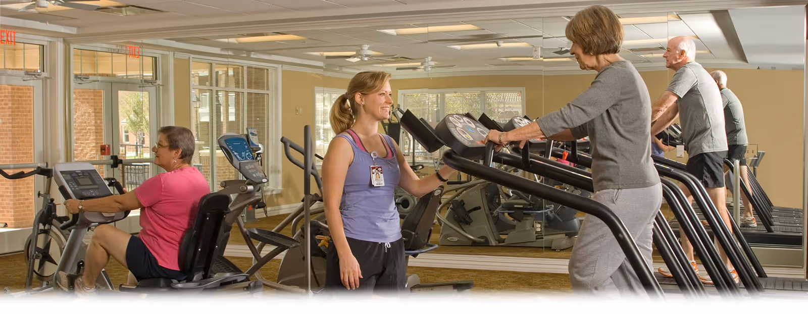 A fitness room in a senior living facility with exercise equipment including treadmills and stationary bikes. An older woman is walking on a treadmill while a younger female staff member in a purple tank top and black pants smiles and assists her. Another older woman is using a stationary bike, and an older man is walking on a treadmill in the background. The room has large mirrors on the wall and windows letting in natural light.