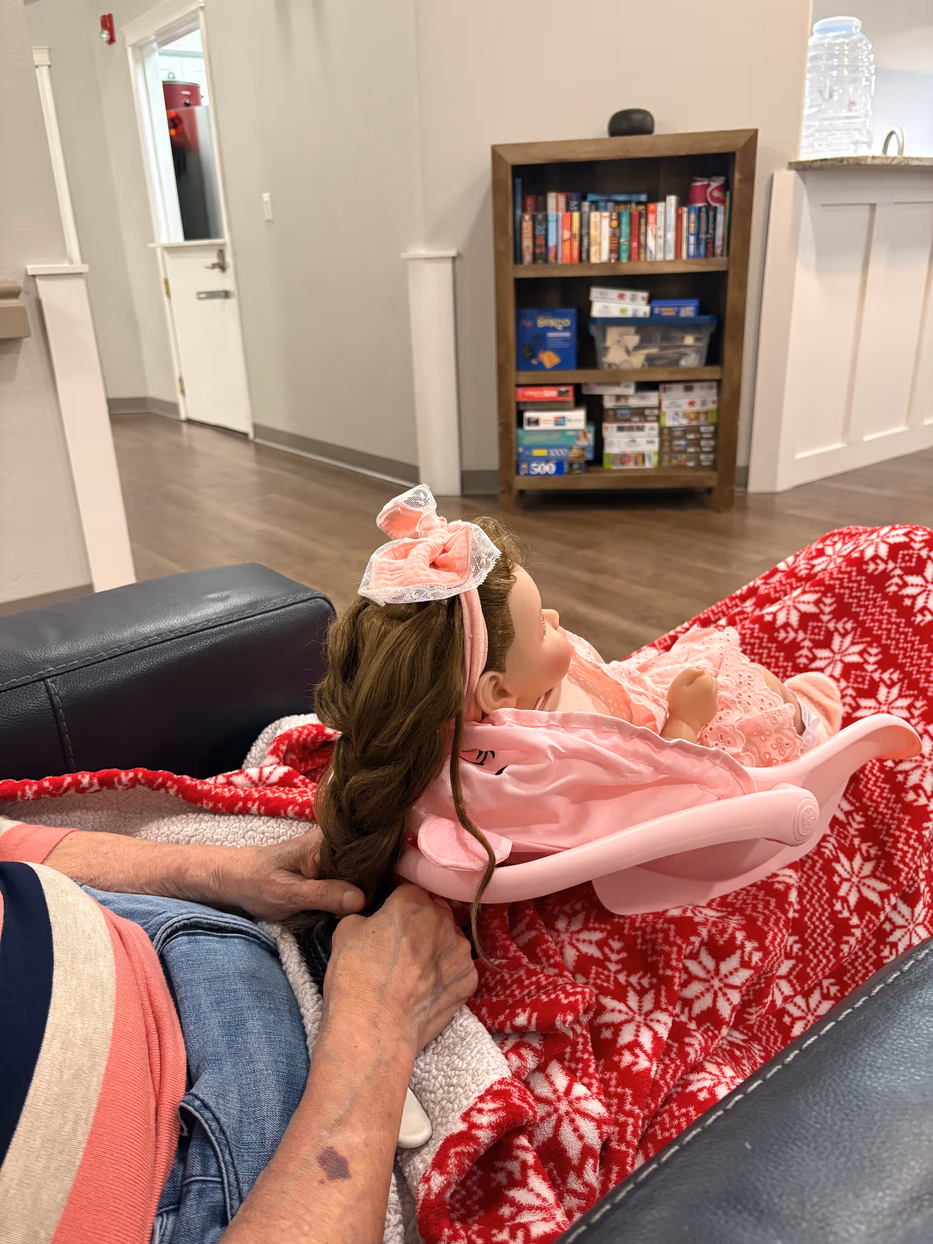 Elderly person sits by a couch with a pink doll in a high chair.