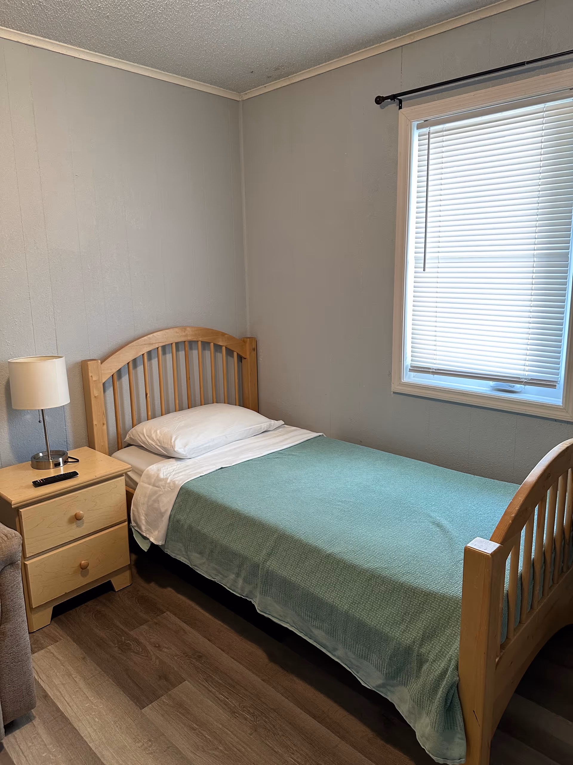 Small wooden single bed with green blanket and nightstand in a simple bedroom.