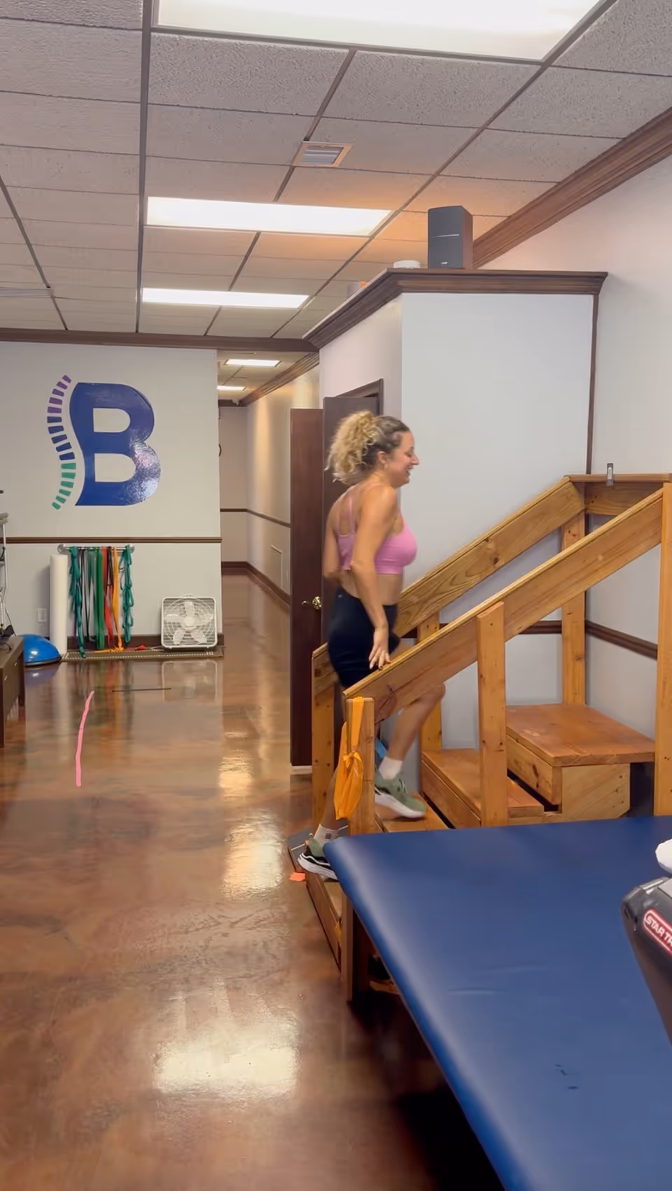A woman in gym attire stands on wooden stairs inside a rehab facility.