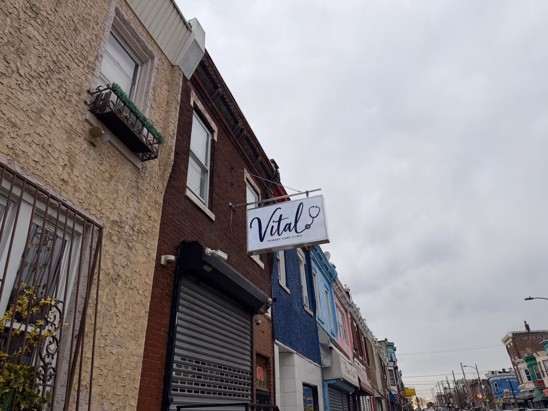Street view of colorful storefronts with a hanging sign reading Vital Primary Care Clinic.