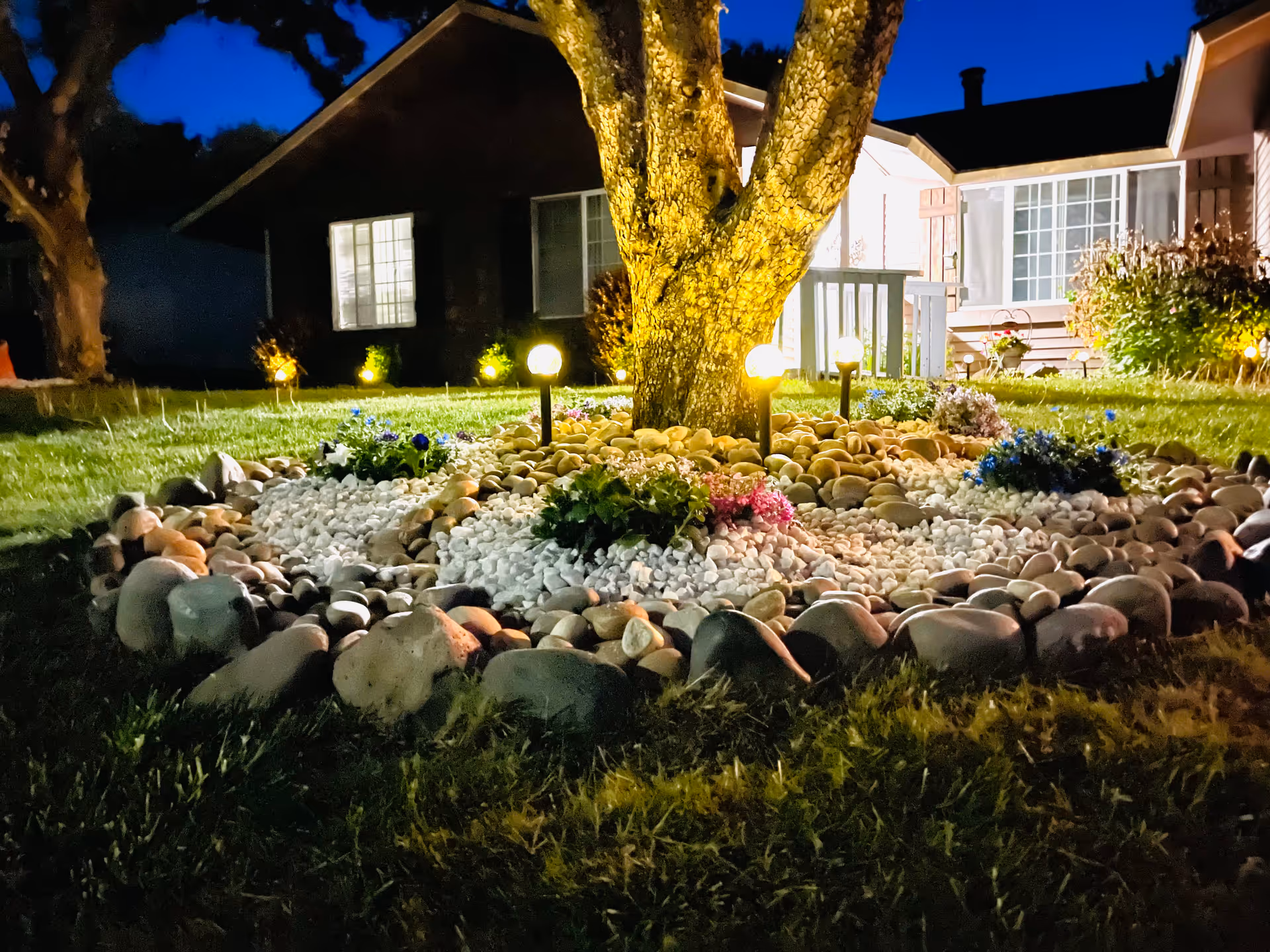 Nighttime exterior of a home with a lit rock garden and lawn