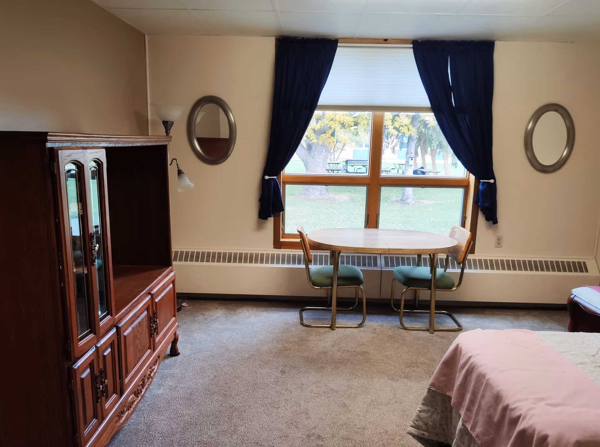 A bedroom with a bed in the foreground, a wooden armoire to the left, and a small round table with two chairs by a large window with dark curtains.
