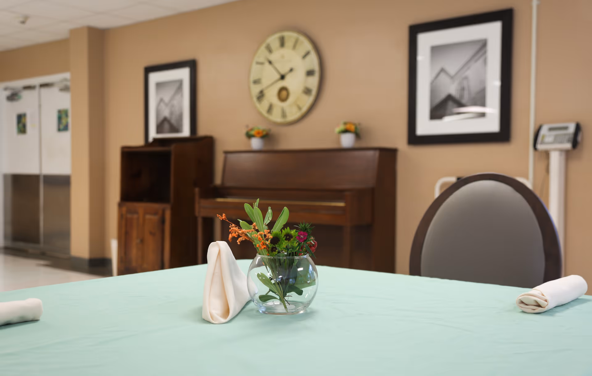 A dining table with a light green tablecloth, a small glass vase with flowers, and two rolled white napkins. In the background, there is a wooden piano, a wooden cabinet, a large round clock on the wall, two framed black and white pictures, and a weighing scale.