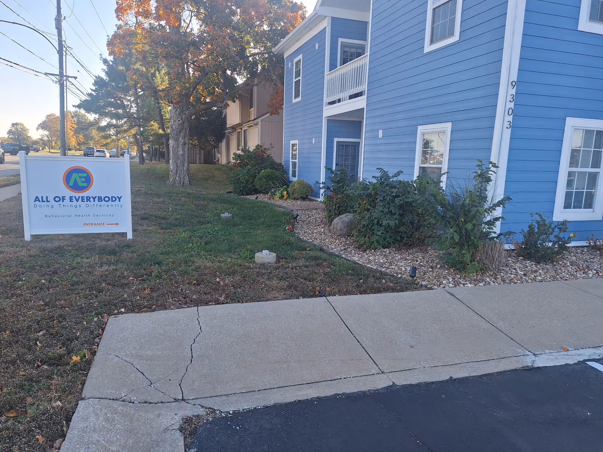 Blue apartment-style building exterior with a sign for All of Everybody in a landscaped yard.
