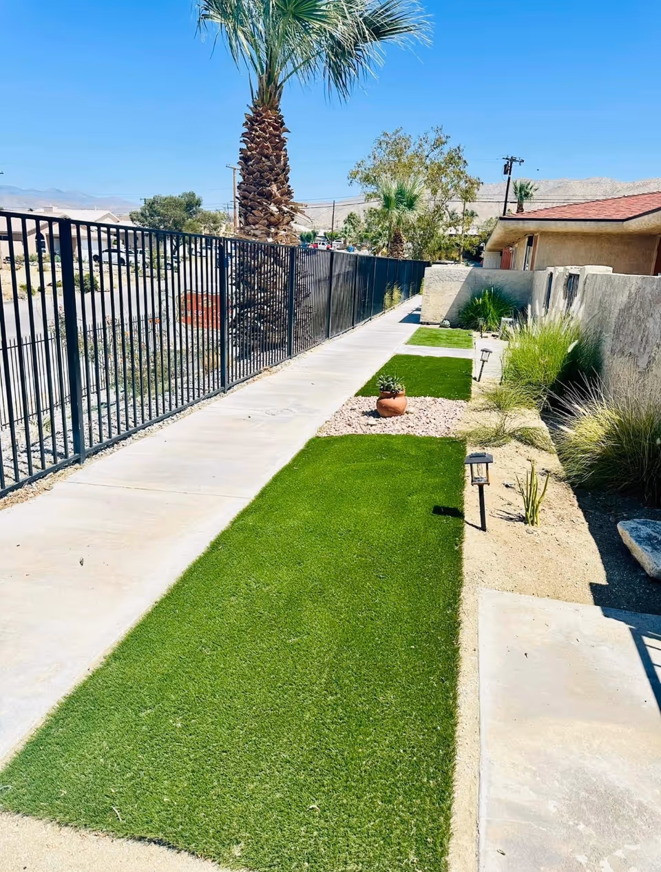 Outdoor walkway at Desert Cove Assisted Living, with a metal fence, artificial turf, and desert landscaping.