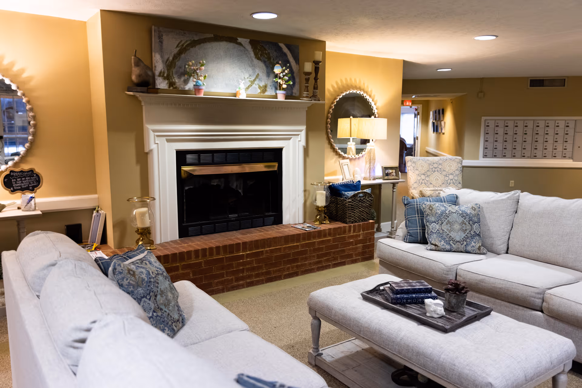 Cozy living room area with light gray sofas adorned with blue patterned pillows, a matching ottoman with a tray holding books and a small plant, a brick fireplace with a white mantel decorated with small potted plants and candles, a round mirror on the wall, a table lamp, and a comfortable armchair in the background.