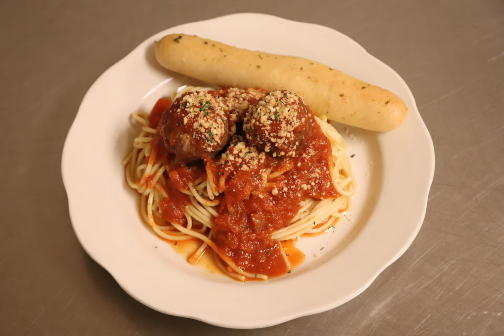 A white plate with spaghetti topped with marinara sauce and three meatballs sprinkled with grated cheese, accompanied by a breadstick on the side.