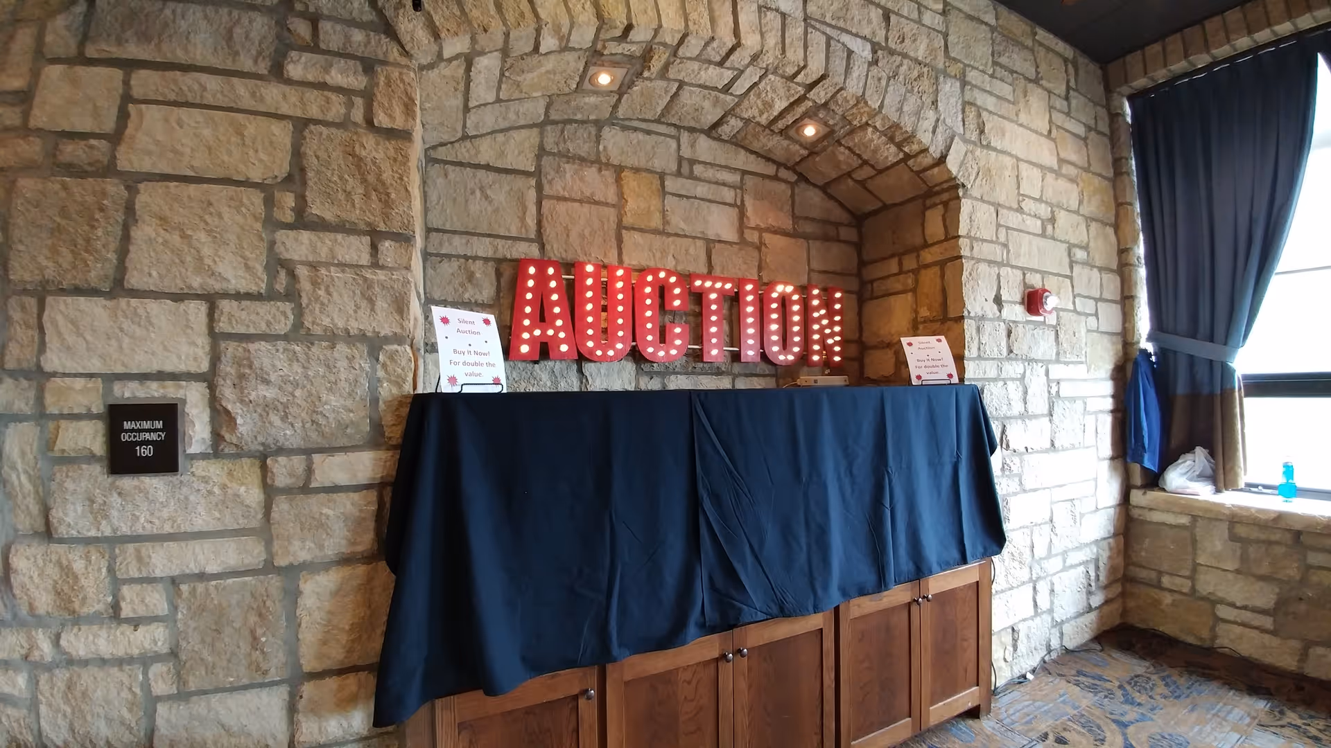 Illuminated AUCTION sign above a blue-draped table in a stone alcove interior.