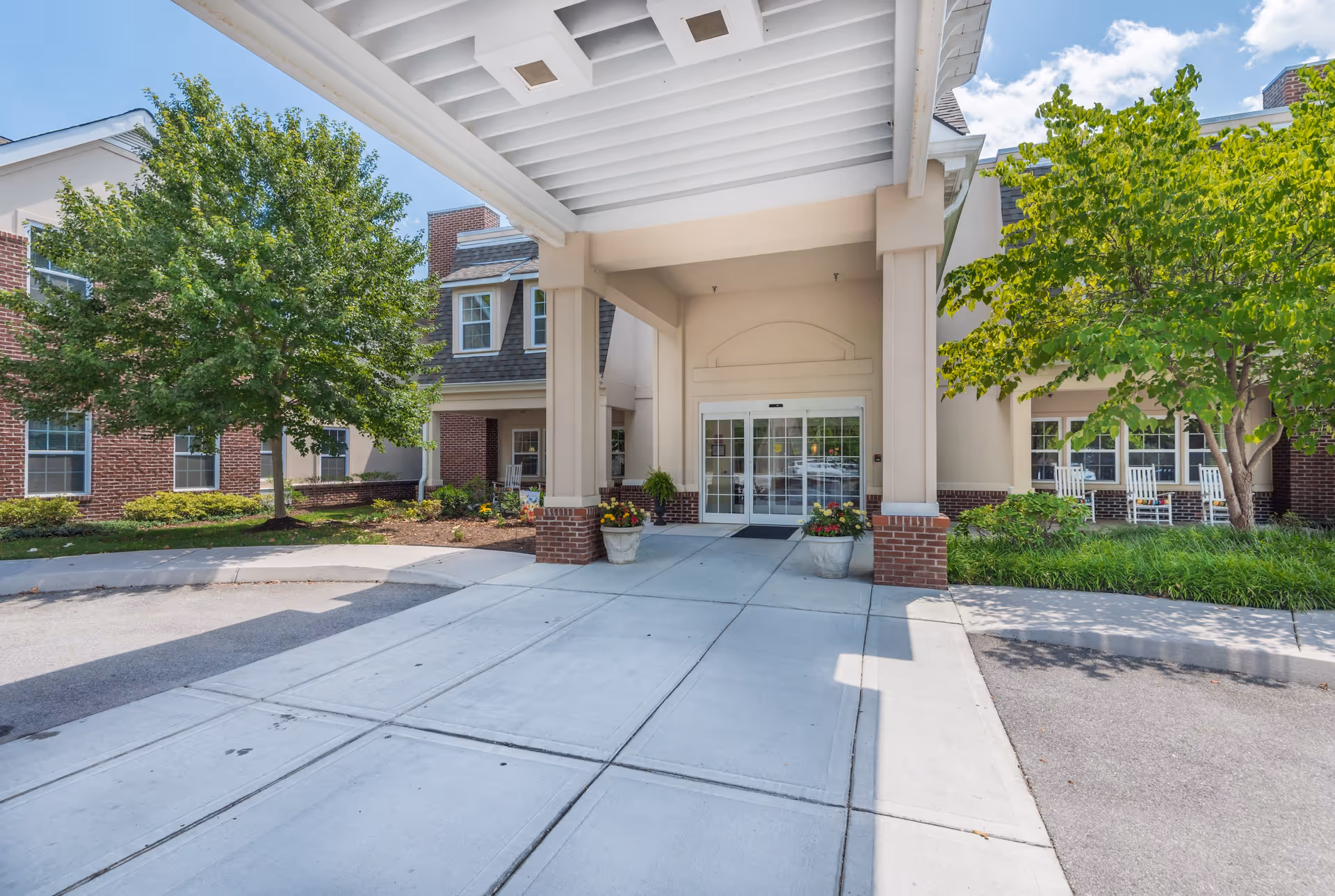 Entrance of Park Place of West Knoxville facility with a covered driveway, two large columns, potted plants with flowers, and a glass double door. The building exterior features brick and beige walls, with green trees and shrubs on either side and rocking chairs visible near the windows.