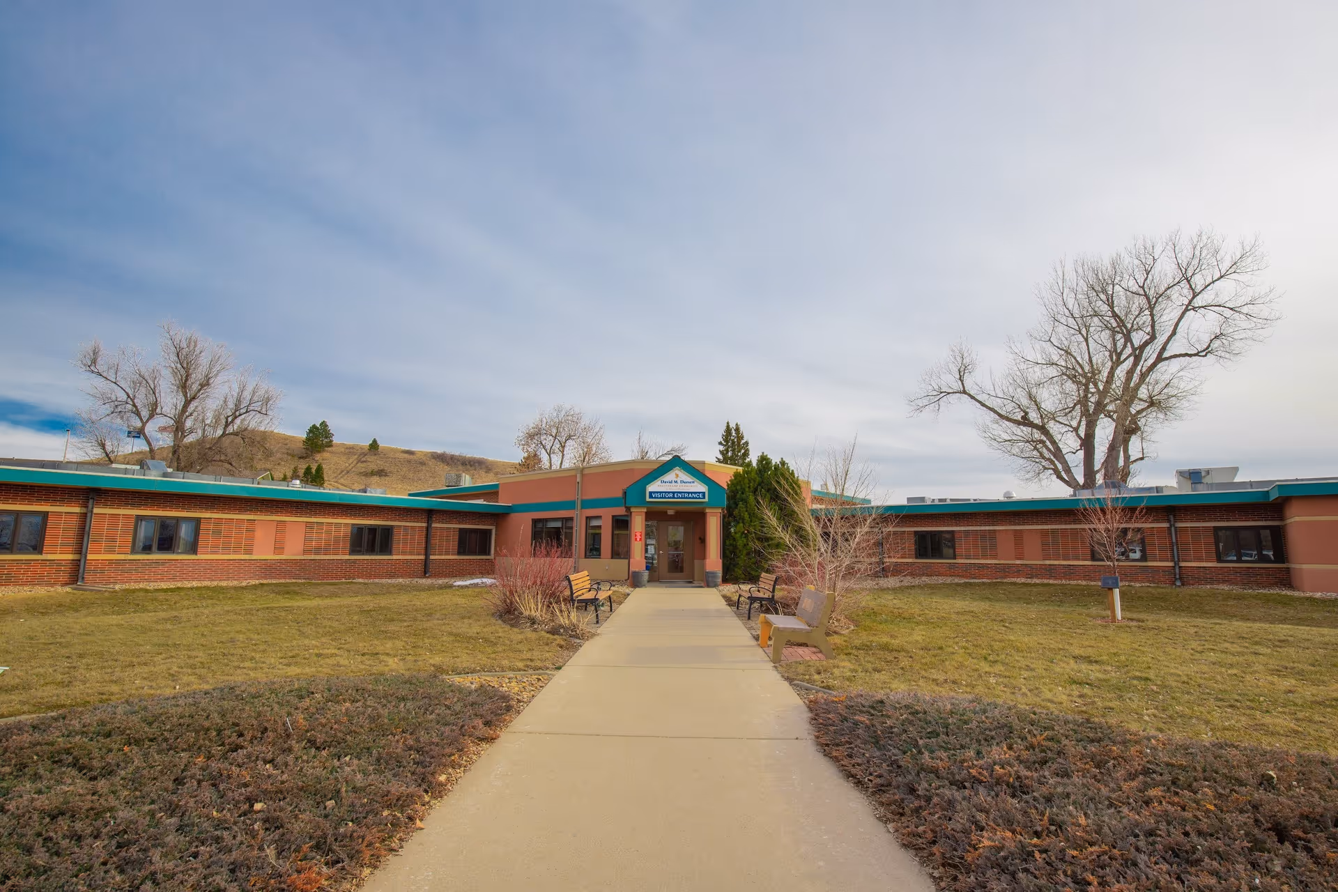 Exterior view of Spearfish Canyon Healthcare building with a concrete walkway leading to the visitor entrance. The building is single-story with brick and brown walls, surrounded by grass, bushes, and leafless trees under a partly cloudy sky.