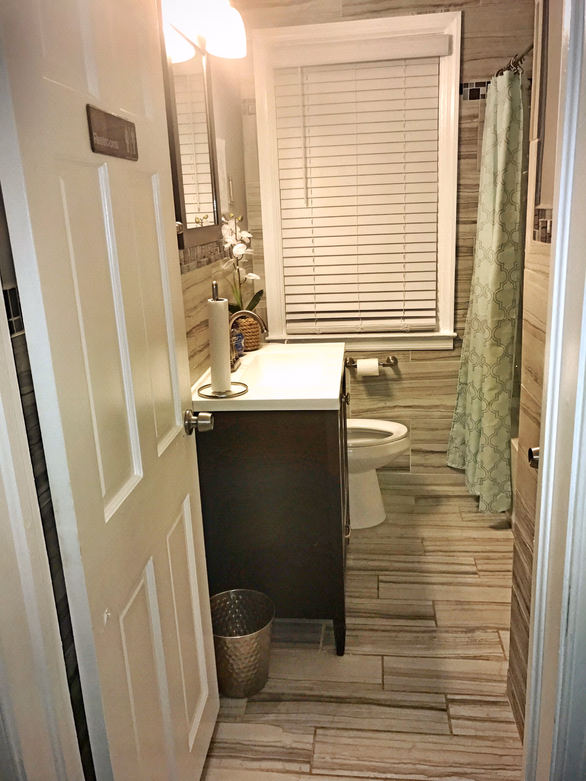Small bathroom with vanity, toilet, shower curtain, window blinds, and tiled walls.