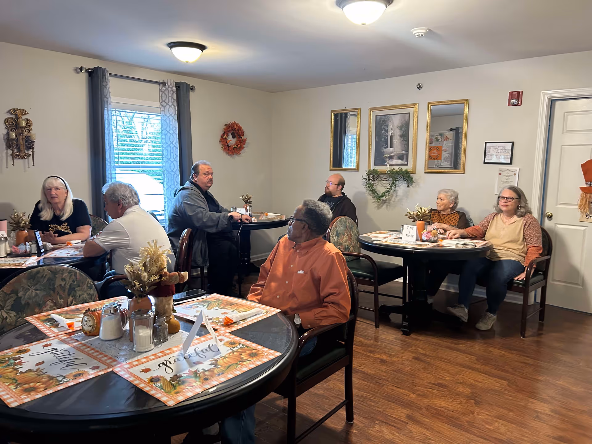 A group of elderly people sitting at round tables in a well-lit room with wooden floors and light-colored walls. The tables have autumn-themed placemats and small decorative centerpieces. The room has a window with curtains, framed pictures and mirrors on the wall, and a door with a fall decoration.