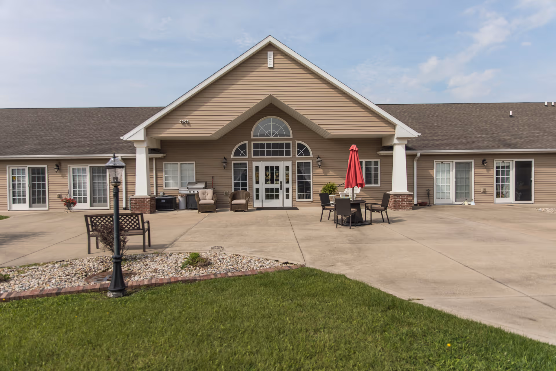 Front exterior of a single-story senior living building with a covered entrance, patio furniture, and a red umbrella.