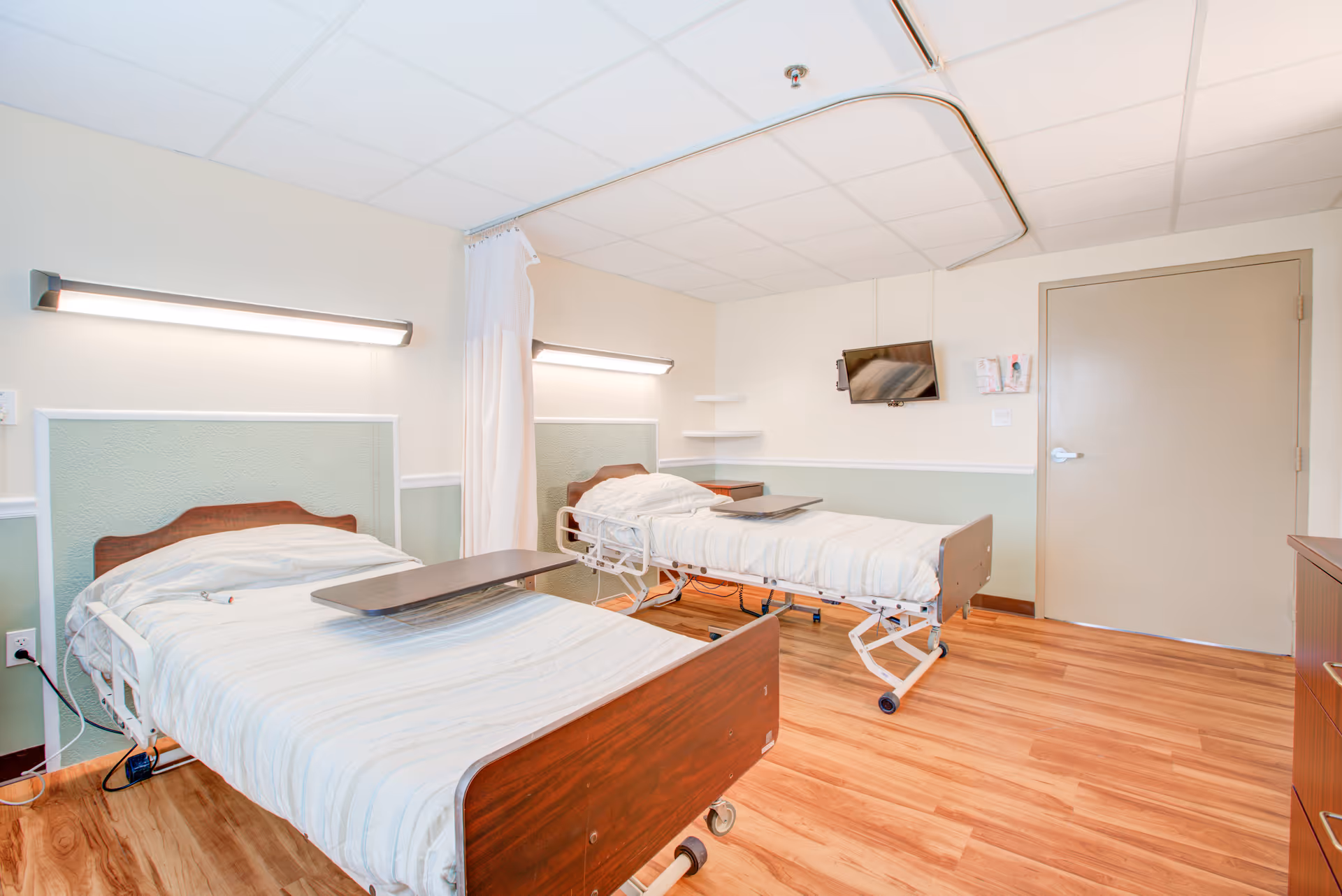 Two hospital beds with white bedding in a bright patient room, separated by a privacy curtain.