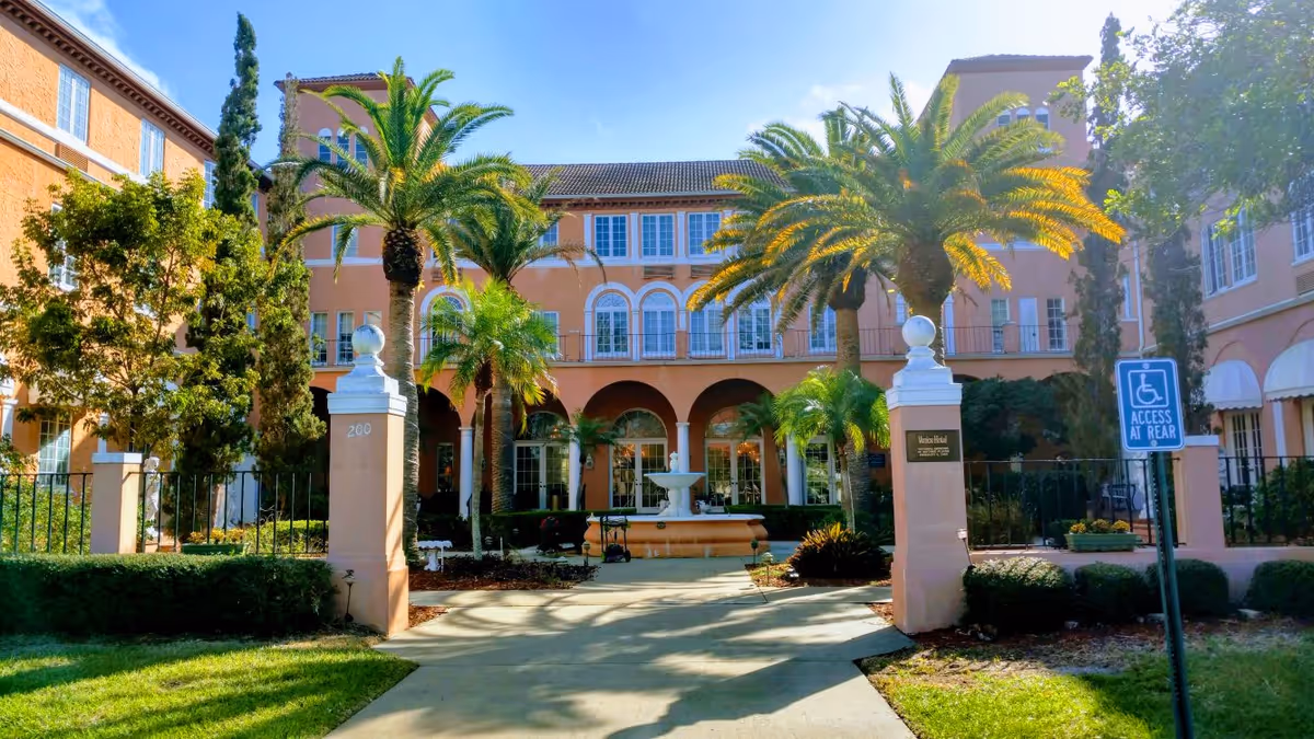 Front entrance of a peach-colored senior living building with palm trees, a central fountain, and a gated driveway.