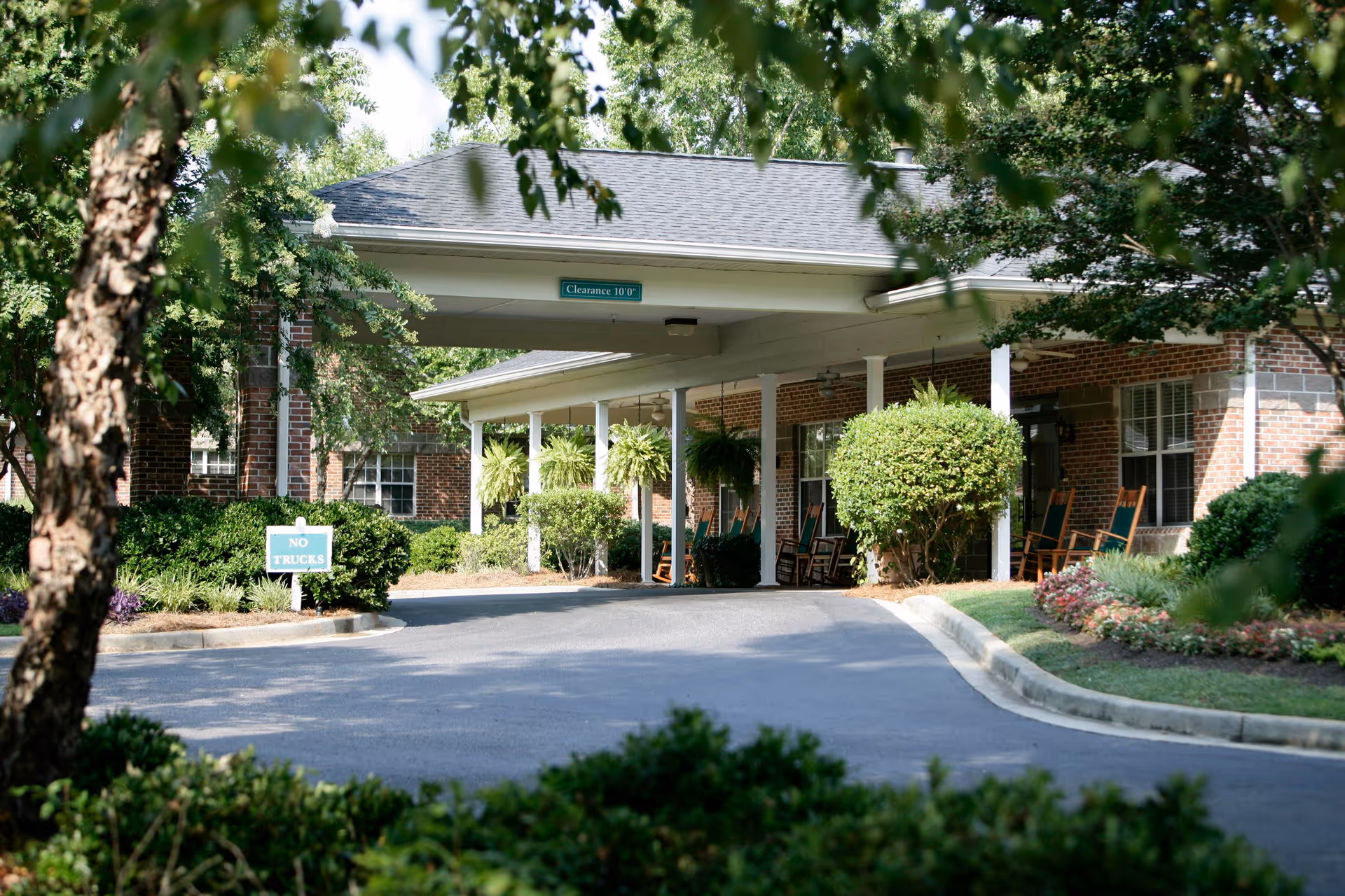 Entrance area of Silver Bluff Grove Senior Living with a covered driveway, brick building, hanging plants, rocking chairs on the porch, and surrounding greenery and trees.