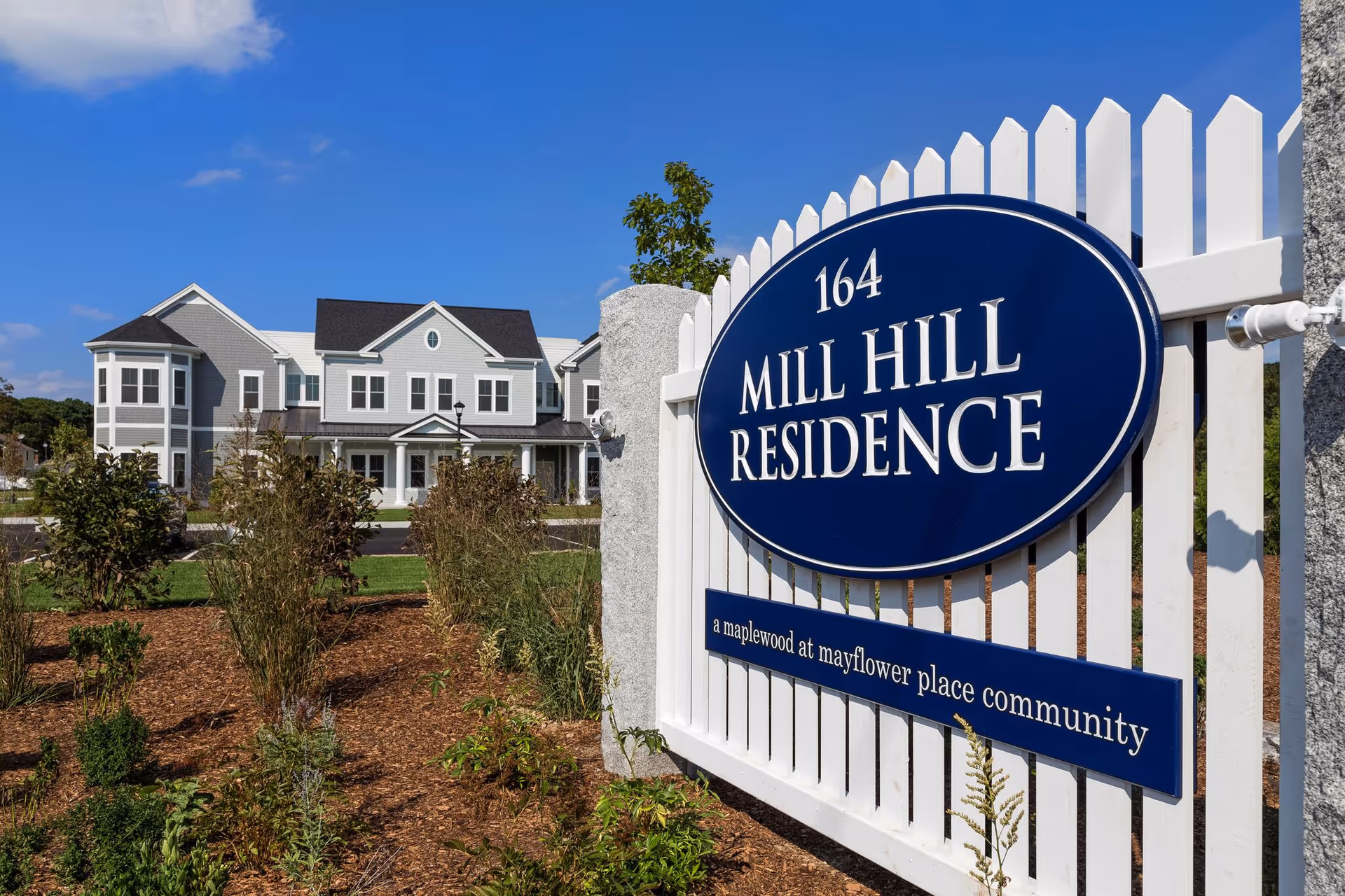 Blue oval Mill Hill Residence sign on white picket fence with building behind.