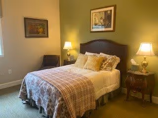 A cozy bedroom featuring a neatly made bed with multiple pillows and a plaid blanket. The room has two bedside tables with lamps, a chair in the corner, and framed artwork on the walls. Soft lighting creates a warm and inviting atmosphere.