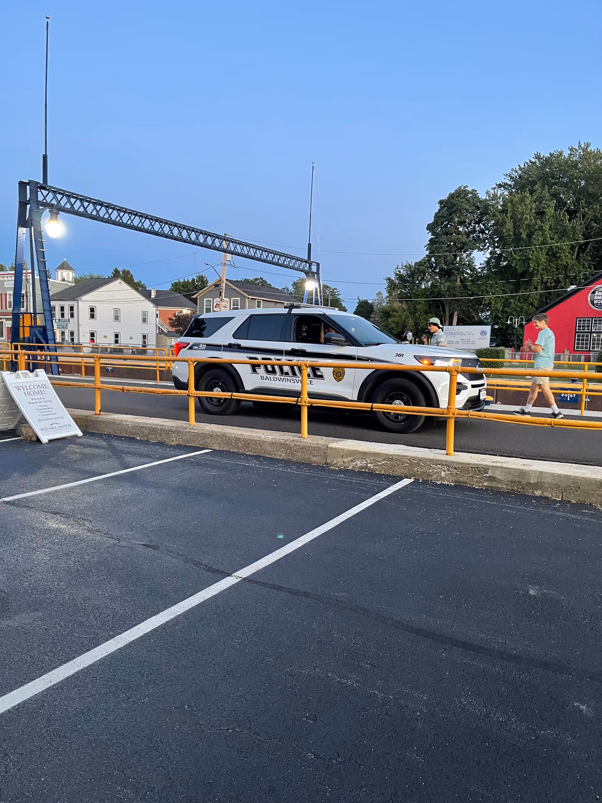 Police SUV with Baldwinsville markings in a parking lot, pedestrians nearby.