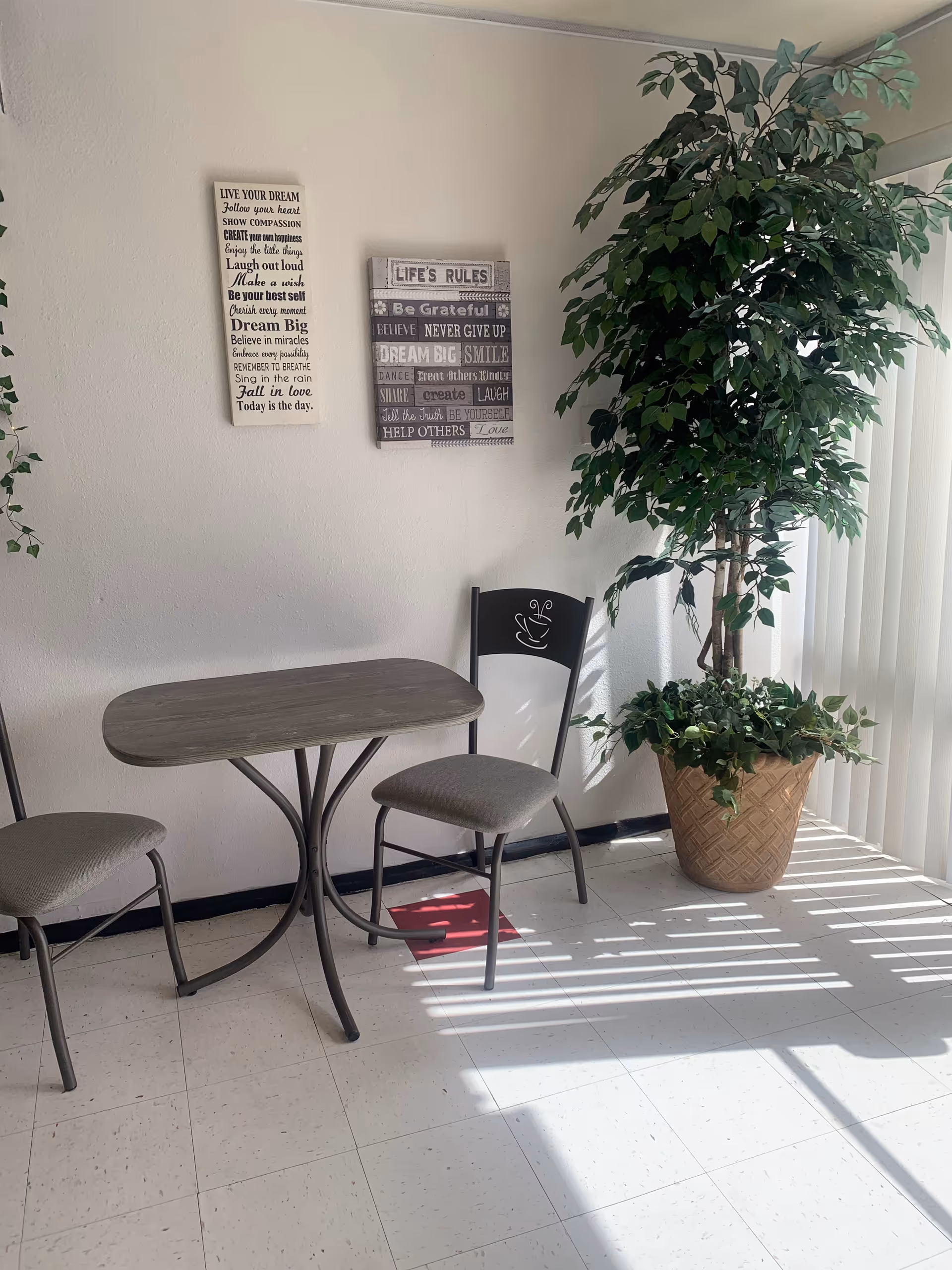 Indoor dining nook with small table, two chairs, plants, and wall art.