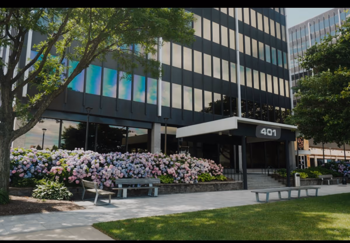 Modern glass-front building exterior with colorful flower beds, benches, and a manicured lawn.