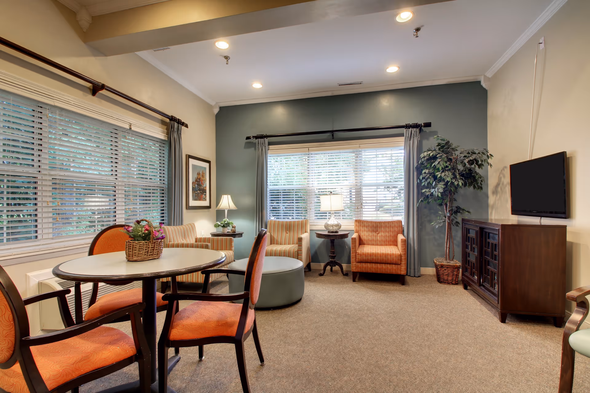 Cozy senior living lounge with a round table and chairs, upholstered armchairs by a large window, TV on a cabinet and a potted plant.