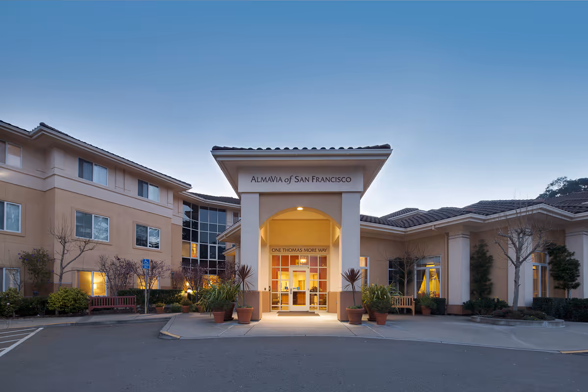 Front entrance of the AlmaVia of San Francisco senior living facility with a covered portico, potted plants, and warm interior lighting at dusk.
